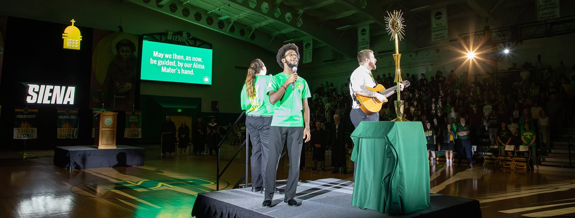 University Celebration moment with performers and production elements in a Siena gymnasium setting.